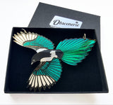 A magpie bird necklace with teal, black, and silver wings, displayed inside an open black presentation box with the brand name 'Obscenerie' visible on the lid.