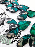 A close-up of multiple magpie necklaces. Bird-shaped charms with teal, black, and silver mirror wings, arranged on a white background with a black-and-white line drawing of a bird.