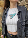 A close-up of a person wearing a teal and silver bird-shaped magpie necklace over a white ribbed crop top and a dark gray denim jacket, standing outdoors against a stone wall.