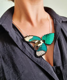 A close-up of a person wearing a silver necklace with a detailed magpie bird pendant featuring bright teal wings and a black and silver head, styled over a dark gray button-down shirt.